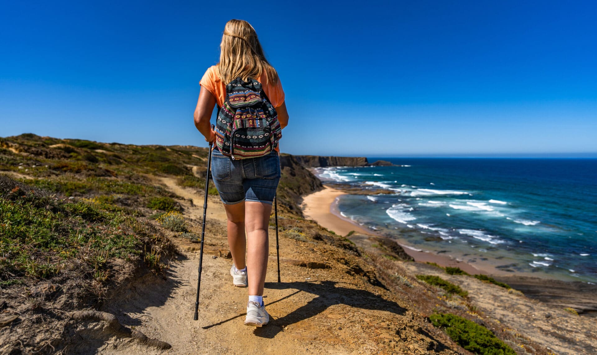 Beautiful mature woman hiker hiking on tourist trail along atlantic ocean in nature park Vicentine Coast in Portugal on sunny summer day. Rear view
